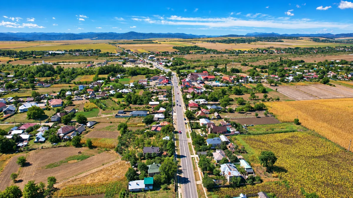 Vedere aeriană asupra comunei Girov, cu drumul principal, case, terenuri agricole și dealuri la orizont.